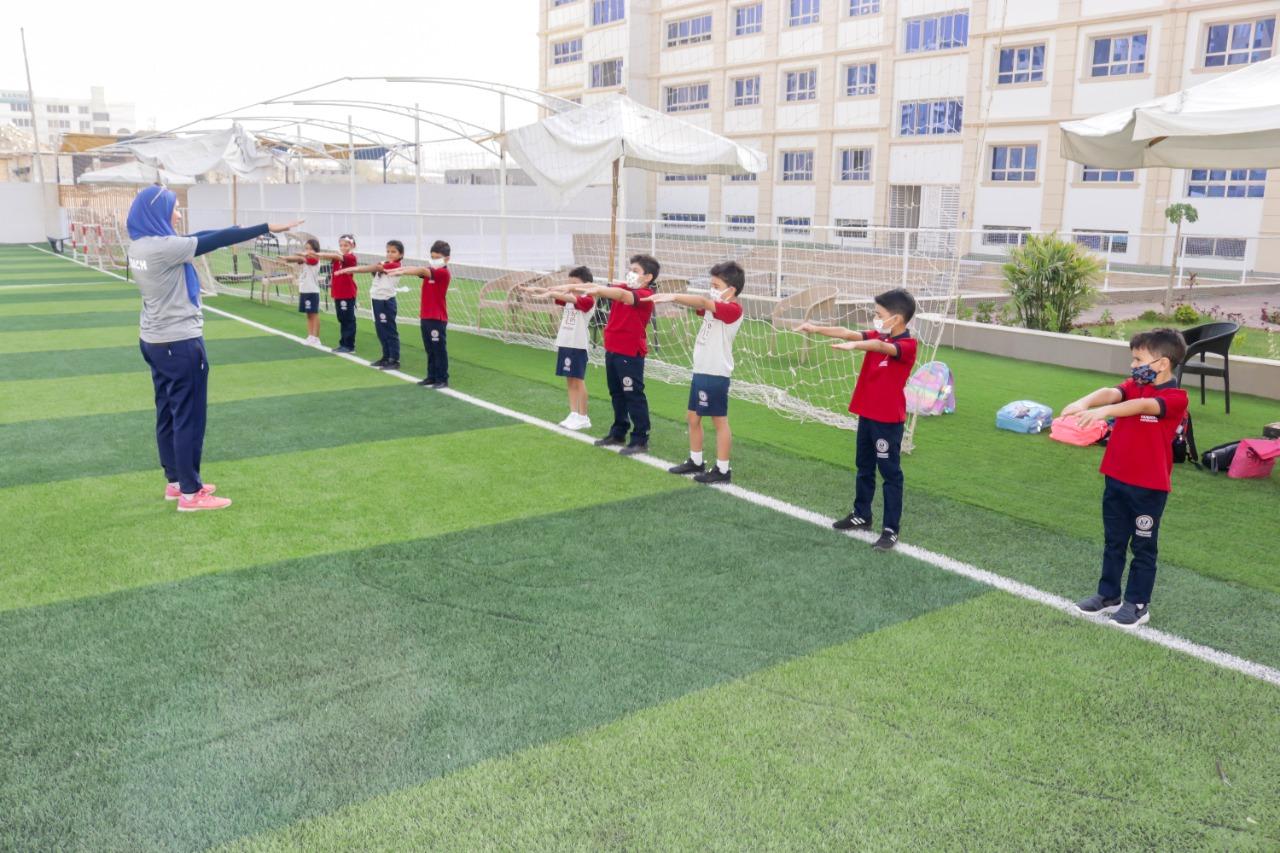 Young students of IVY STEM International School participating in a physical education class or sports practice on the school's sports field. They are seen performing stretching and exercise routines under the guidance of a coach or teacher. The sports field features artificial grass, a net for sports like soccer, and surrounding buildings and tents, indicating a well-equipped facility for sports and recreational activities.